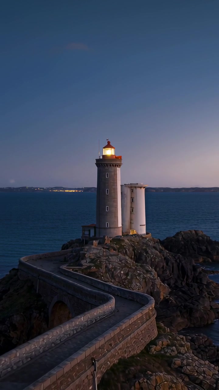 Video verticale: Faro guida che proietta un fascio di luce mentre il crepuscolo si intensifica sul mare, con una piccola torre e una strada rialzata.