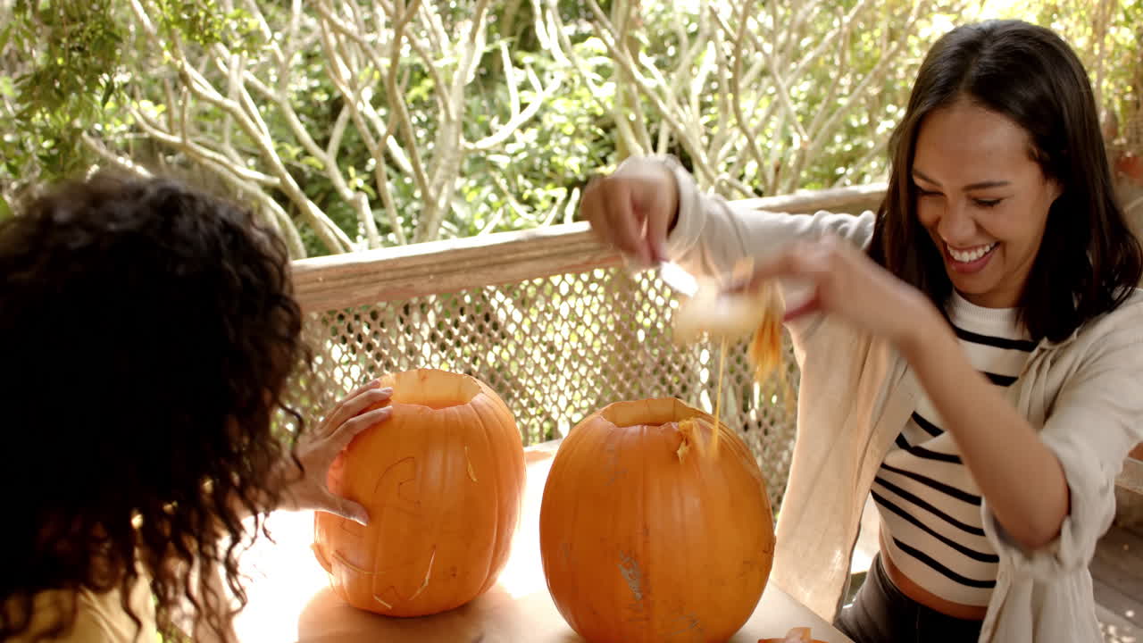 Halloween time, smiling multiracial female friends carving pumpkins on a porch