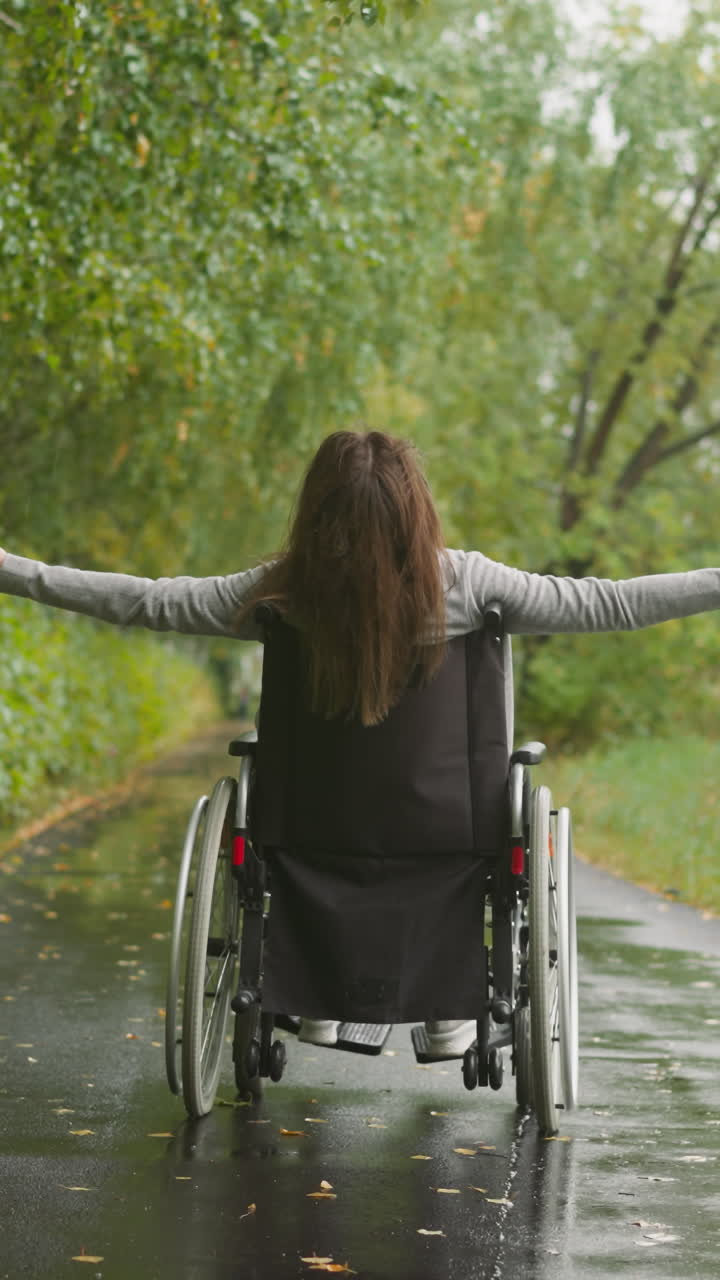Woman with spinal cord injury stretches arms to sides enjoying walk in park on rainy day. Female recovers after accident resting in sanatorium backside view