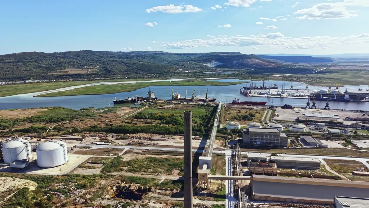 Área industrial perto do rio com navios de carga atracados e terreno verde ao fundo durante um dia ensolarado