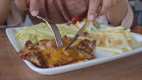 close-up of a person cutting grilled chicken with a knife and fork, served with pasta and french fries