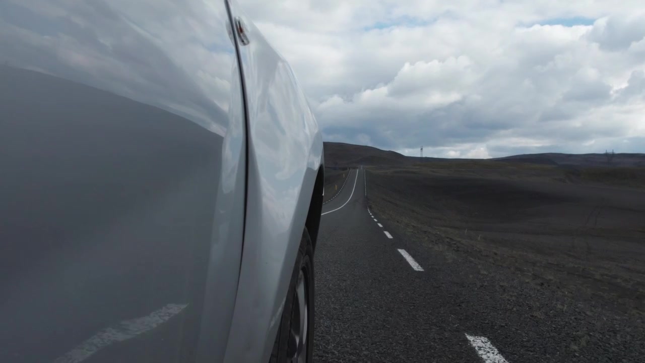 Un'inquadratura laterale di un'auto che guida lungo una strada remota attraverso un deserto in Islanda. Il veicolo si muove attraverso il vasto paesaggio brullo.