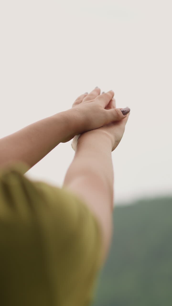 Woman catches rain drops and spreads onto hands against distant green sloppy hill at eco resort on nasty day backside view slow motion