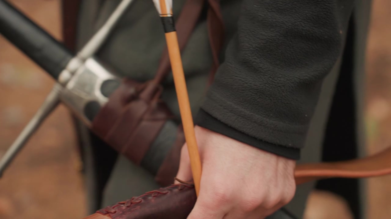Primo piano di una mano che impugna il riser dell'arco e tende la corda su un arco ricurvo in legno, con freccia incoccata, flettenti curvi in tensione, posizionamento stabile delle dita e dettagli esterni nitidi in una calda luce naturale.