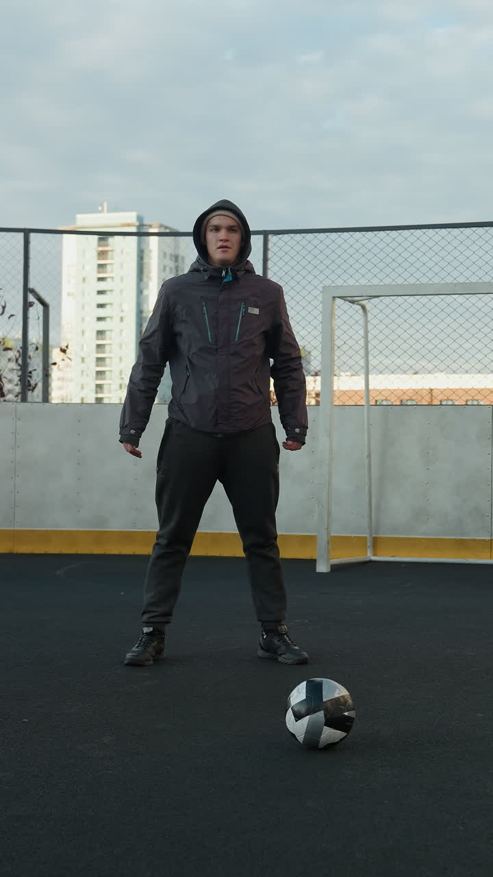 Athlete performing squats with hands clasped in preparation for exercise on outdoor sports ground with soccer ball in foreground, surrounded by urban high-rise buildings and goal post