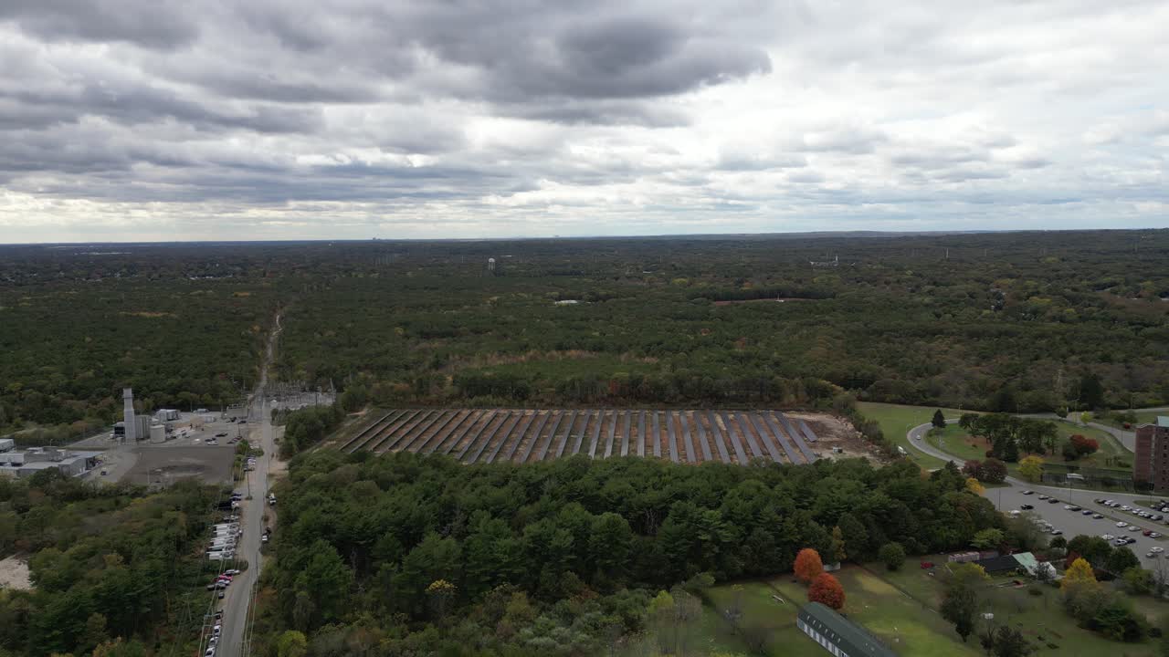 Aerial drone pass over a large solar farm in Brentwood, Suffolk County, NY, showing long rows of solar panels surrounded by autumn trees and open land under soft overcast daylight conditions