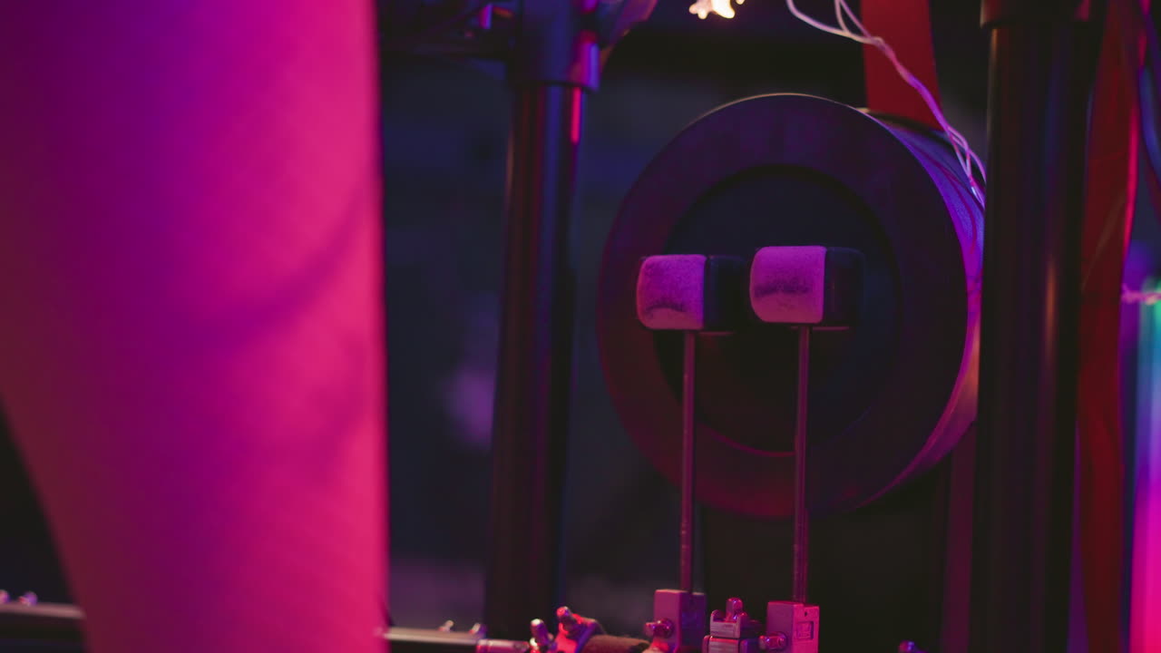 Close up of leg in fishnet pantyhose pressing twin beaters against kick drum pedal in studio setting with pink and purple neon lighting and surrounding wires during music practice session