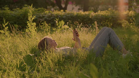 young woman lying on grassy field in warm sunlight, browsing her phone with serene, relaxed expression, tall green grass surrounds her, with blurred building in background