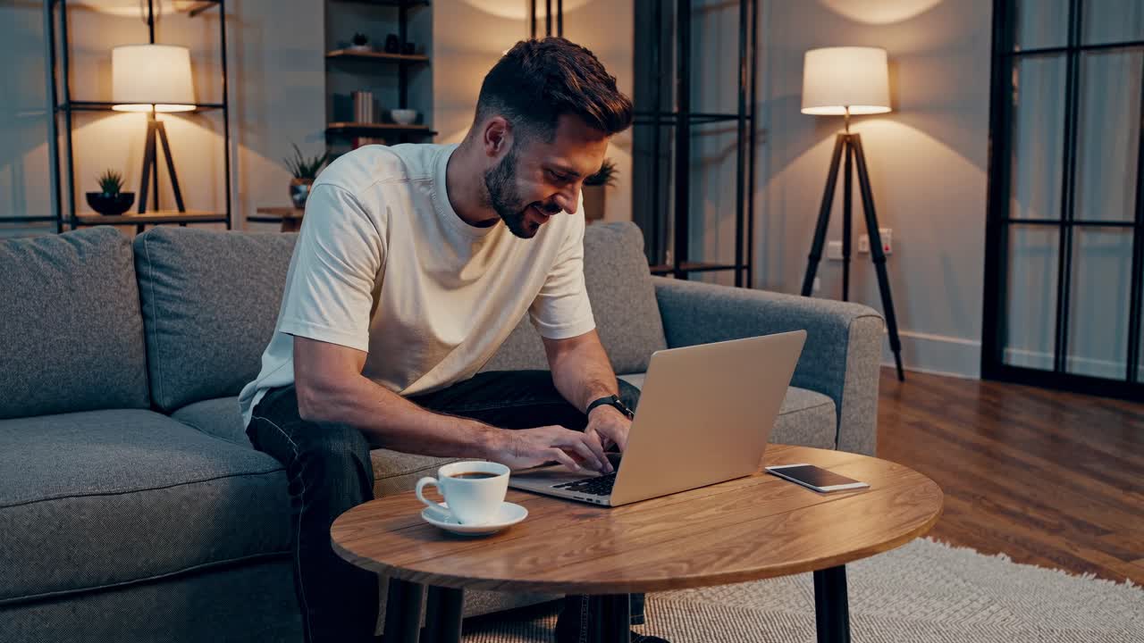 A man works on a laptop in a cozy living room. The video captures a side angle