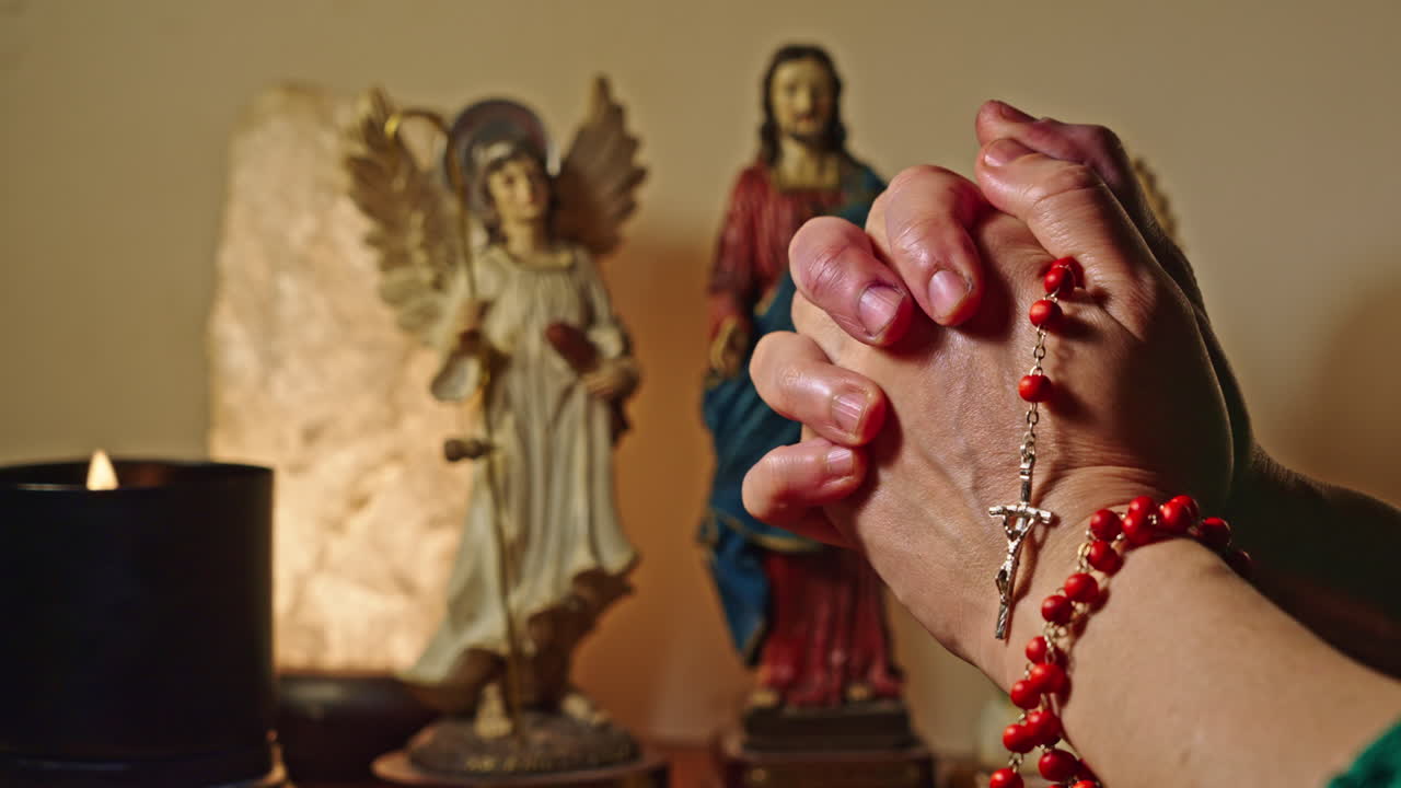 Mani in preghiera che tengono un rosario rosso con statue religiose sullo sfondo