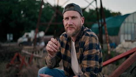 Portrait-of-a-happy-guy-farmer-in-a-cap-who-holds-a-spikelet-in-his-teeth-and-poses-on-a-combine-among-the-greenhouses-on-the-farm