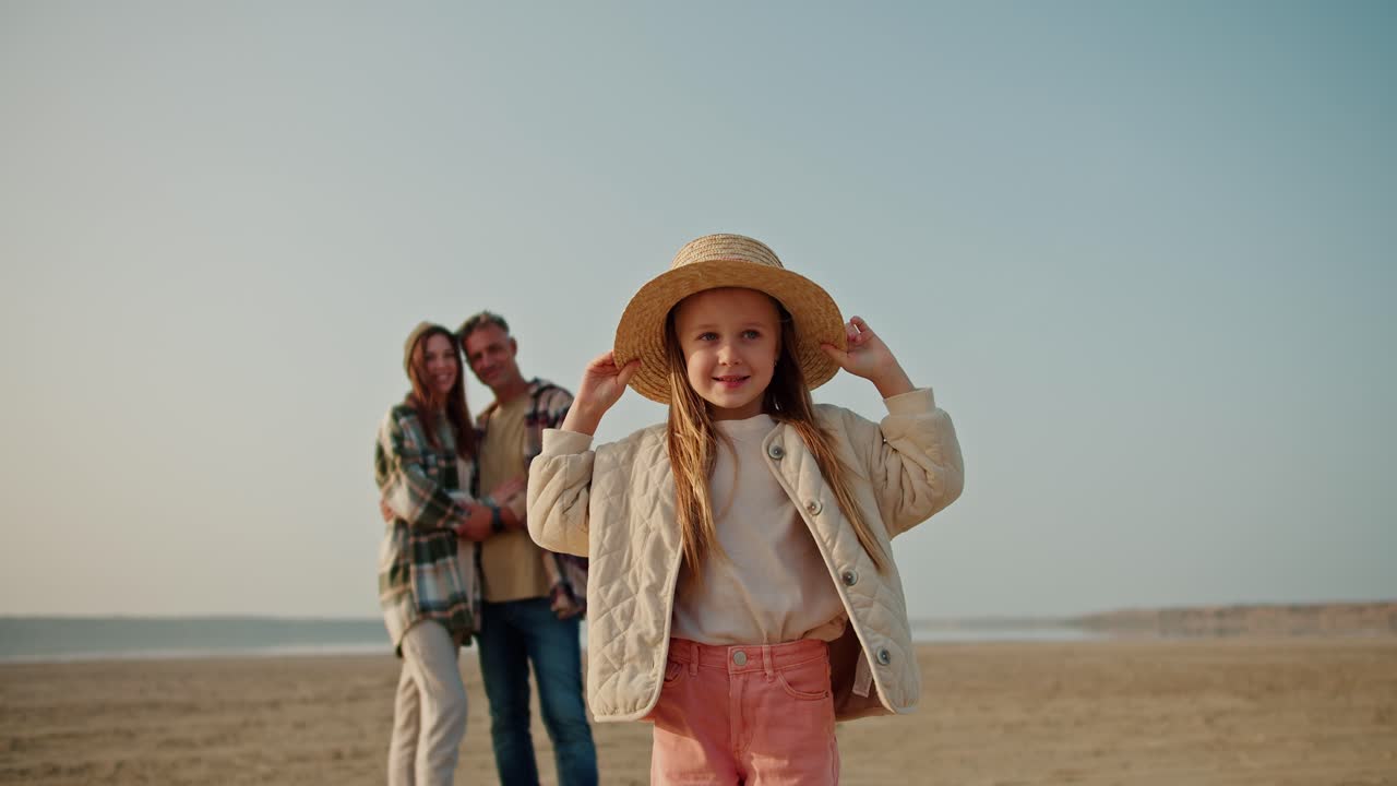 Portrait of a happy little blonde girl in a white jacket, pink pants and a Straw hat who stands and rests during her picnic with her parents, a brunette man with gray hair and a brunette girl in a Green checkered shirt on a deserted seashore in summer