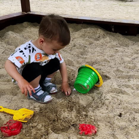 little caucasian kid sitting in the sandbox poured water from his bucket. baby boy touches the wet sand looking pensively away.
