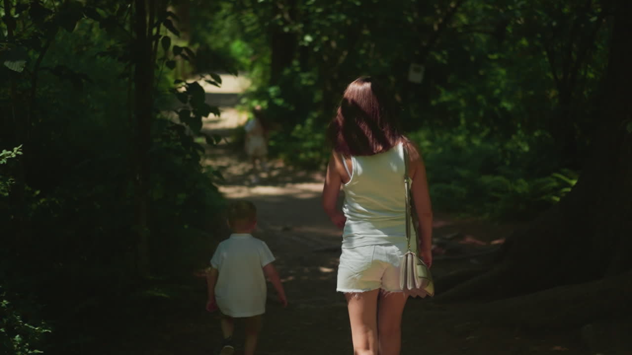 молодая женщина с маленьким мальчиком идет по тенистой дороге через лес в медленном движении. мать с маленьким сыном наслаждается выходными в парке природы. семейный отпуск