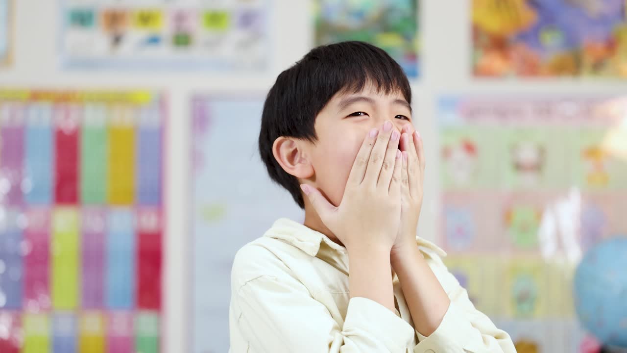 明るい教室で、アジア系の少年が顔に手を当てて、ショックと興奮を表しています。