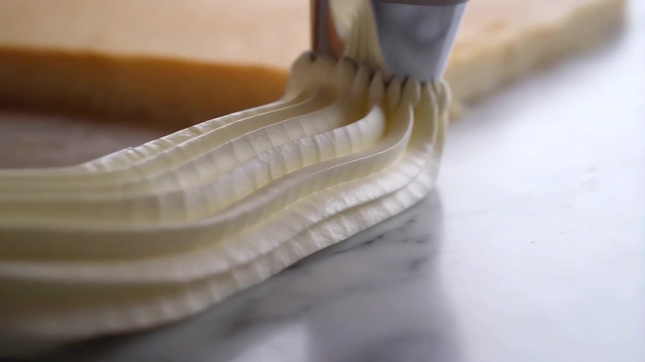Close macro view of glossy buttercream flowing from reflective metal nozzle onto rectangular biscuit on marble countertop, showing ridged folds, scalloped peaks, creamy shine and confection detail