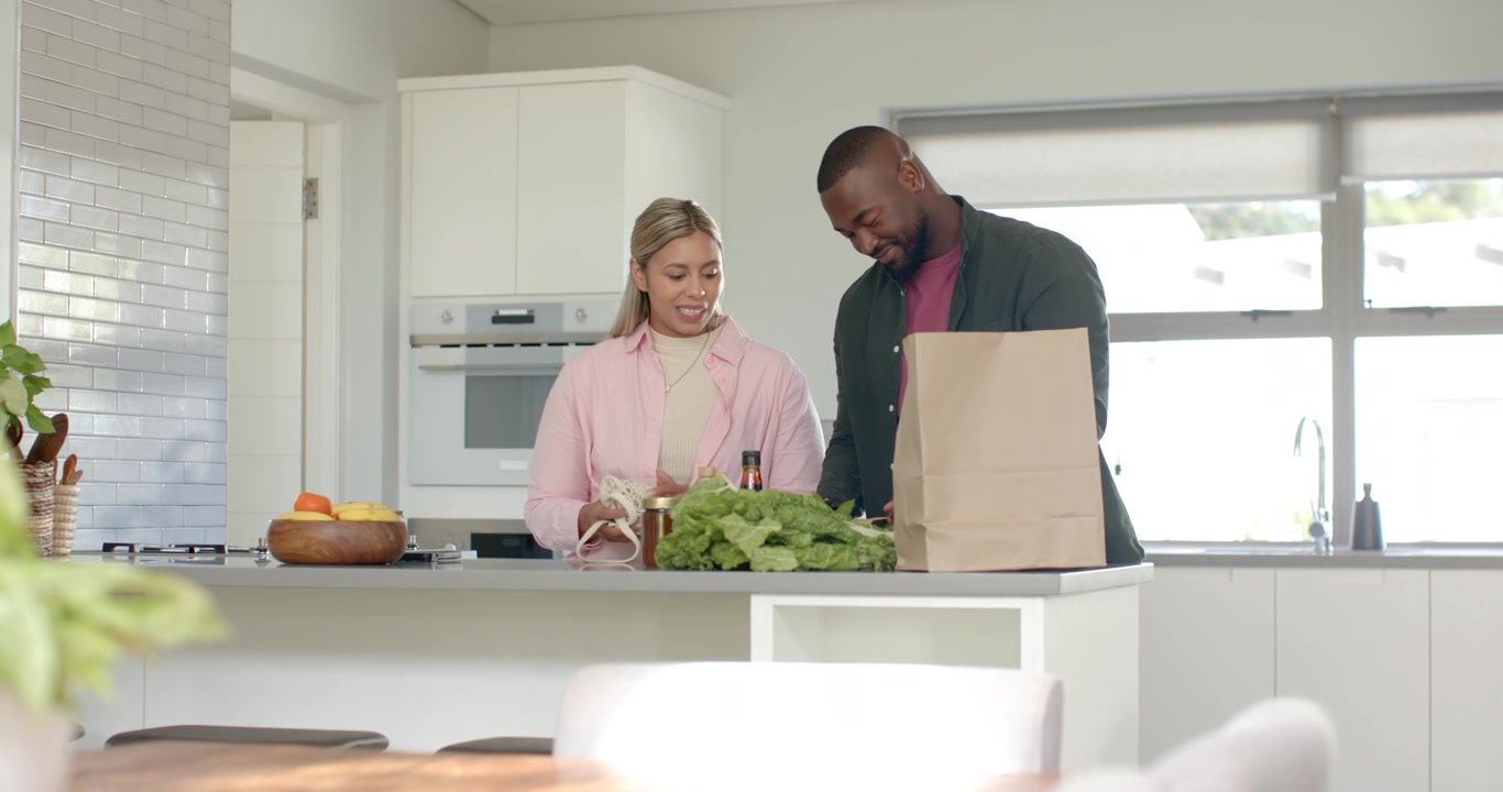 Par packar upp matvaror vid köksön, öppnar en papperspåse och arrangerar grönsaker för middagen.