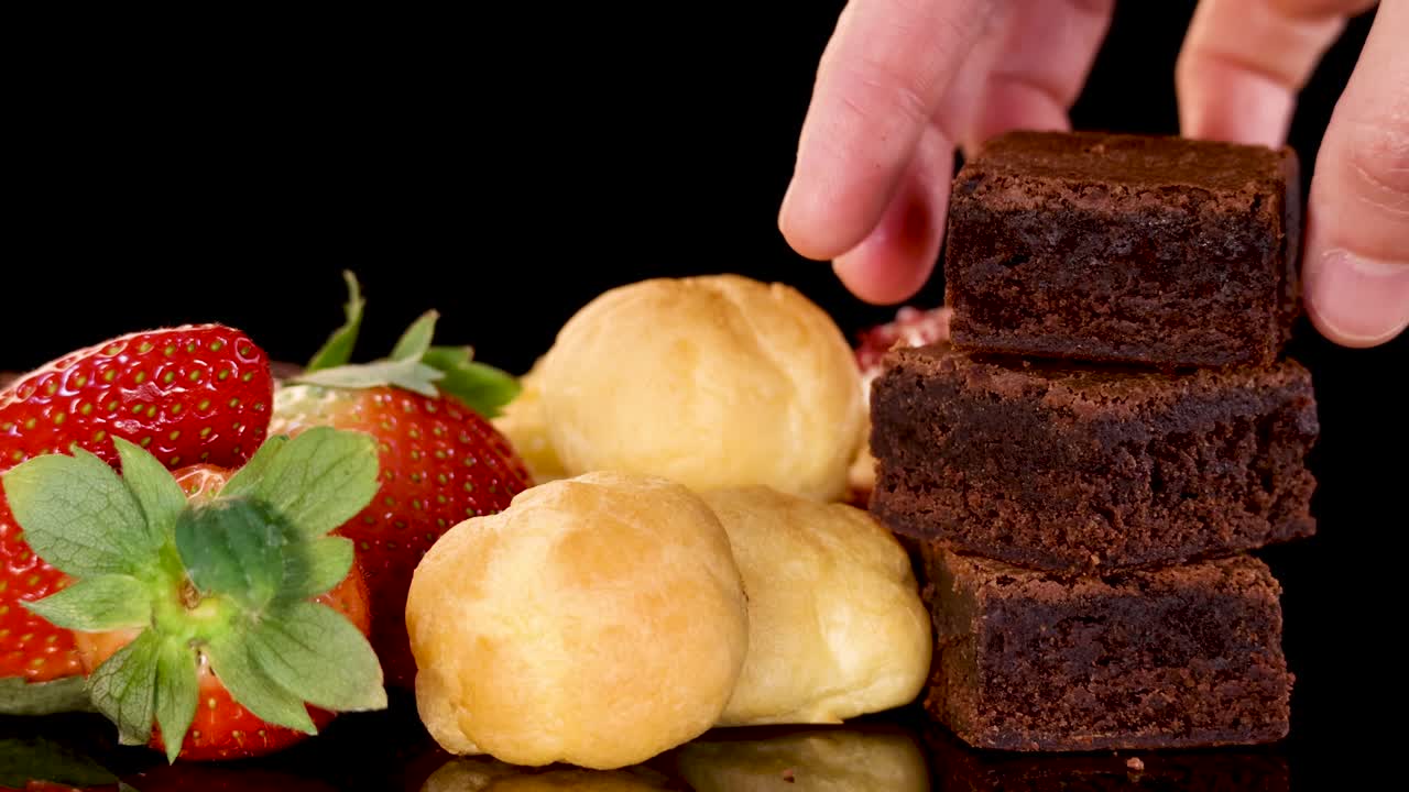 A hand selects a chocolate brownie from a neatly arranged assortment of brownies, cream puffs, and strawberries on a reflective black surface under bright studio lighting