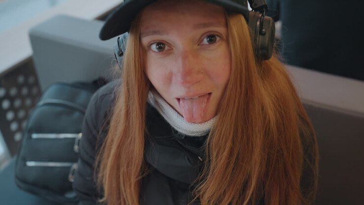 Donna dai capelli rossi con cuffie in caffè, pendolare seduta vicino alla finestra con zaino, si guarda intorno pensierosa, sorrisi sottili e guance lentigginose, avvolta in cappotto e berretto, tensione da viaggio urbano, ruoli