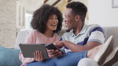 happy african american couple using tablet and smartphone in living room