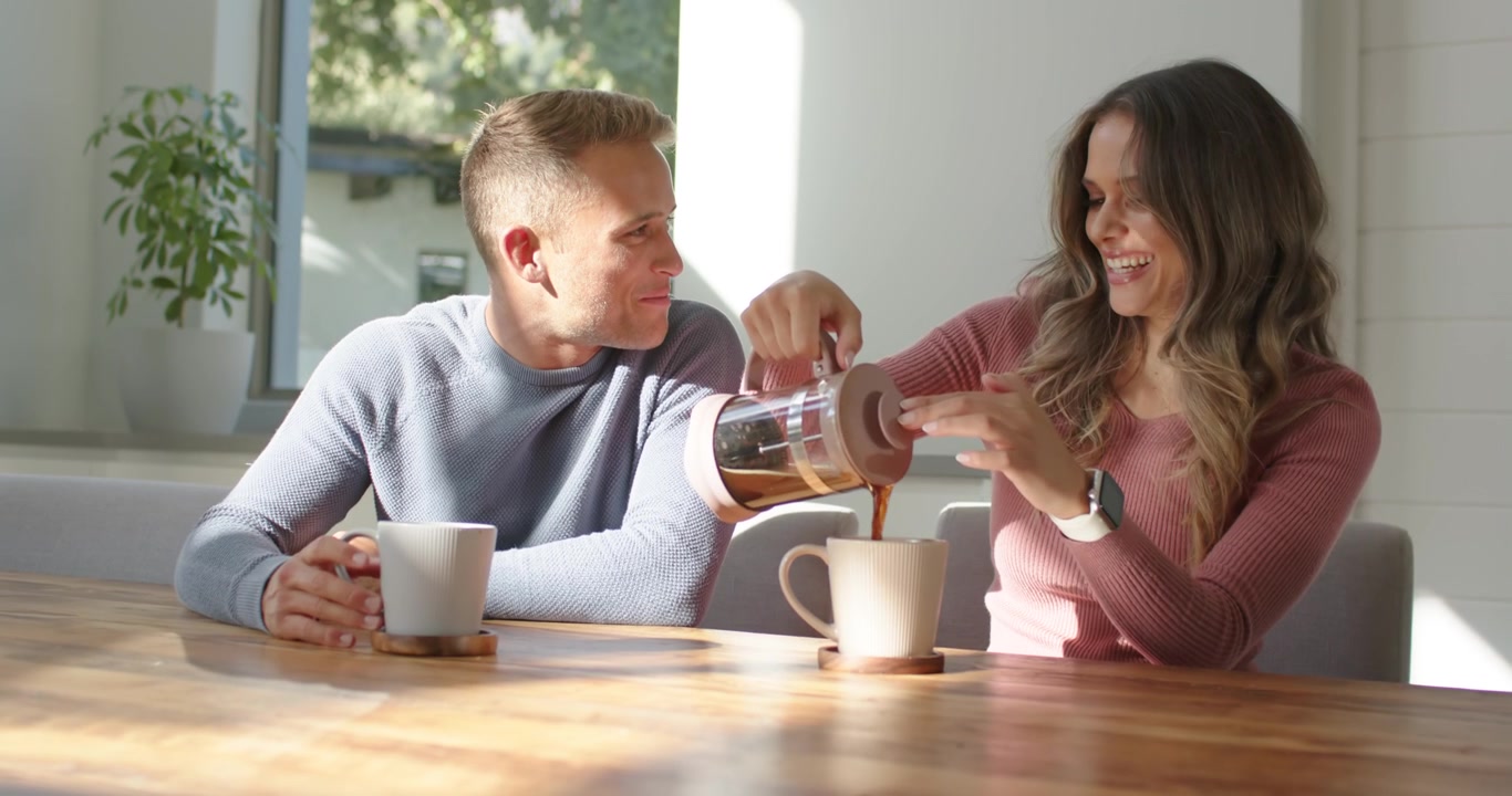 Pareja diversa sentada en una mesa, la mujer tomando una cafetera de prensa francesa y sirviendo café en una taza, compartiendo.