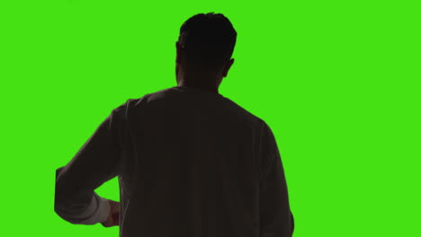 rear view of man answering call on mobile phone standing against green screen studio background with low key lighting