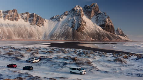 vestrahorn mountain in iceland, aerial wilderness winter landscape of snowy coast lands, tracking tourists van moving away from cars of visitors, rocky mountain peaks in background