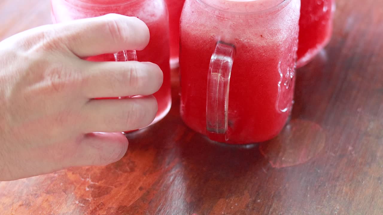 Hands reach for vibrant watermelon juice in mason jars on a wooden table. Bright natural lighting enhances the refreshing scene