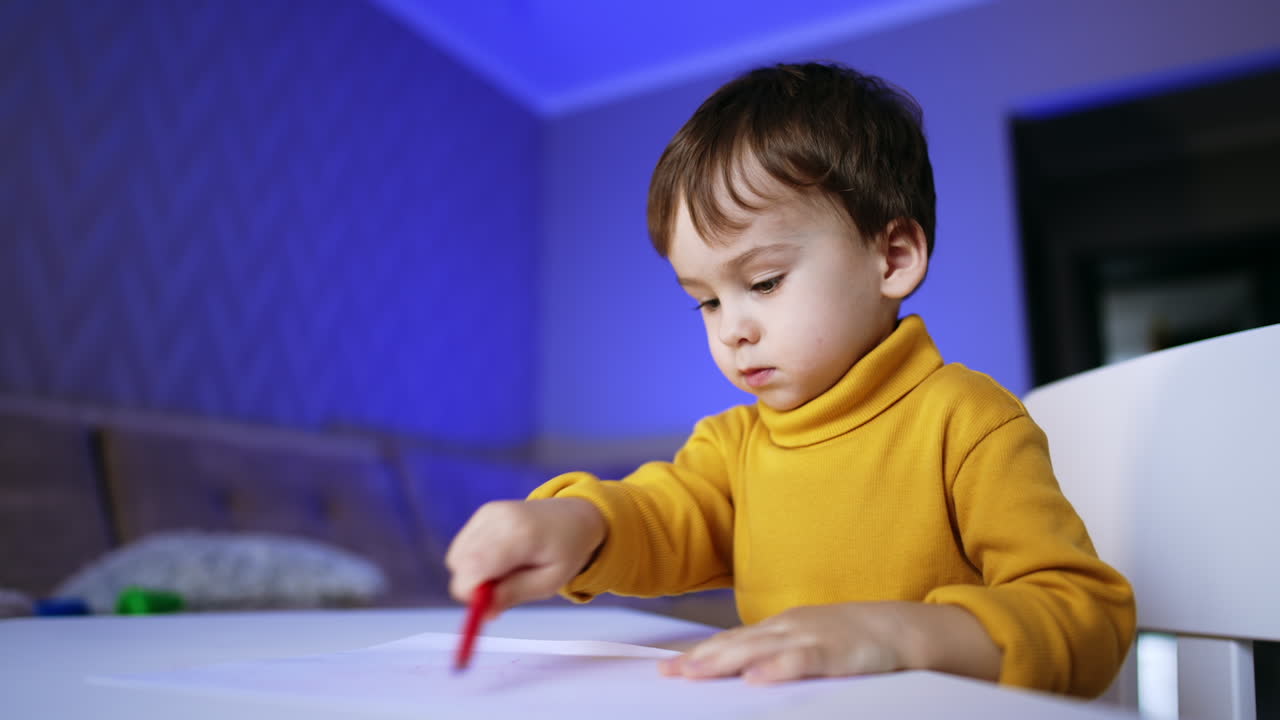Serious toddler boy sitting at desk drawing with felt-pen. Infant child puts the pen and stands up.