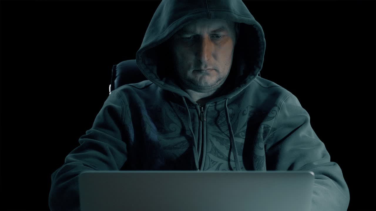 Hacker in a hoody sweatshirt is sitting at table at night and coding. Hacker man in internet. Hacker write and glue sticker to laptop. Techology criminal system designer.