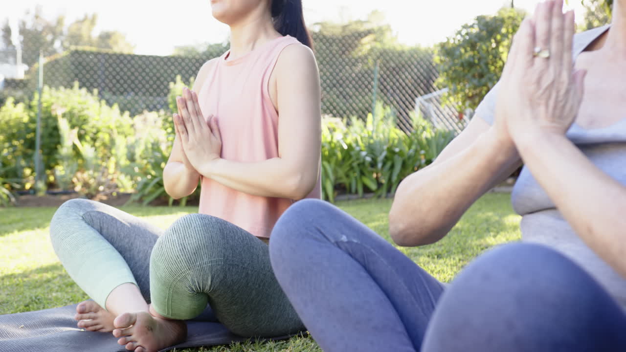 Practicing yoga, Asian grandmother and granddaughter sitting cross-legged on mats in garden
