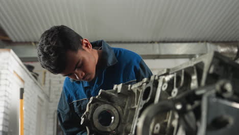 mecánico en uniforme azul inspeccionando componentes del motor en un taller mecánico con vista parcial de otra persona que lo observa, centrado en el trabajo de reparación y la maquinaria