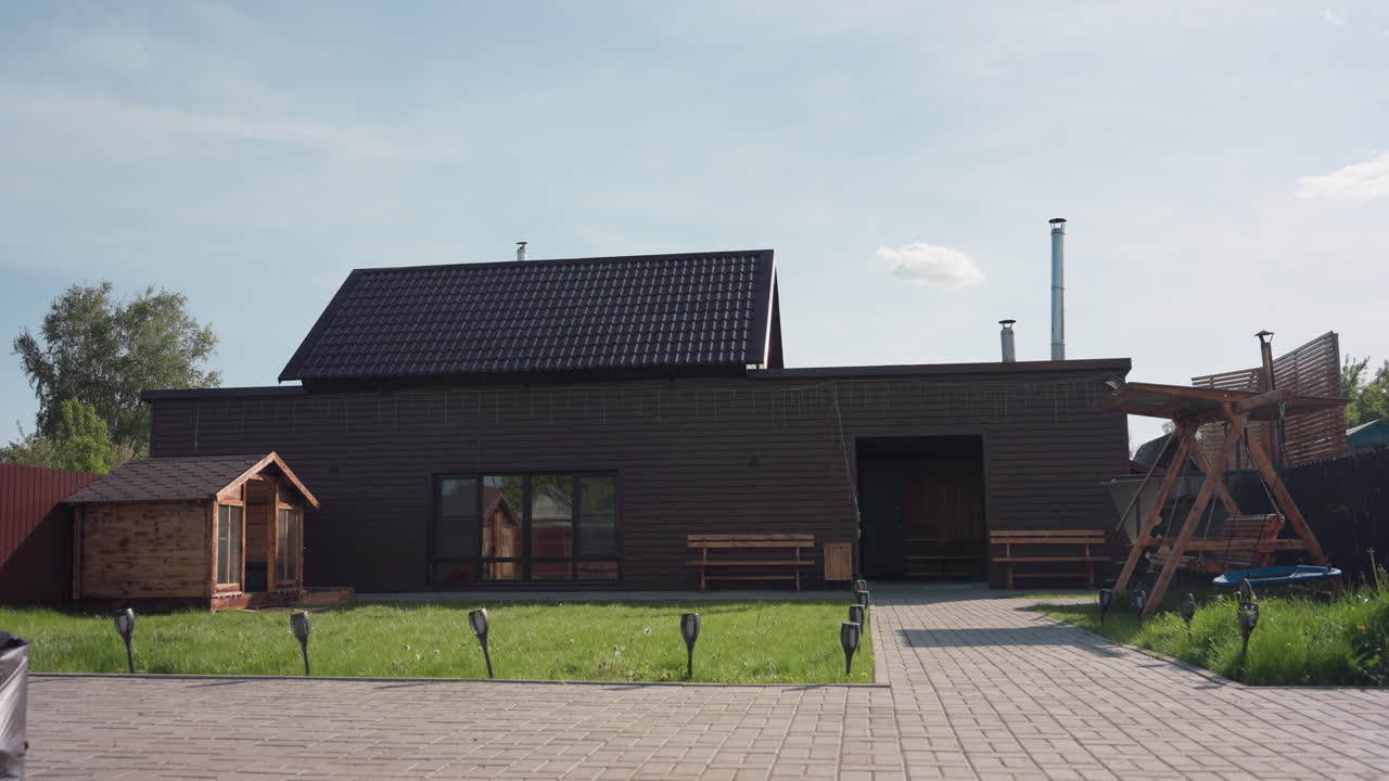Brown wooden house in sunlit fenced compound featuring wooden swing, benches, small cabin, and lush green lawn swaying gently in breeze under clear sky with soft clouds and peaceful backyard
