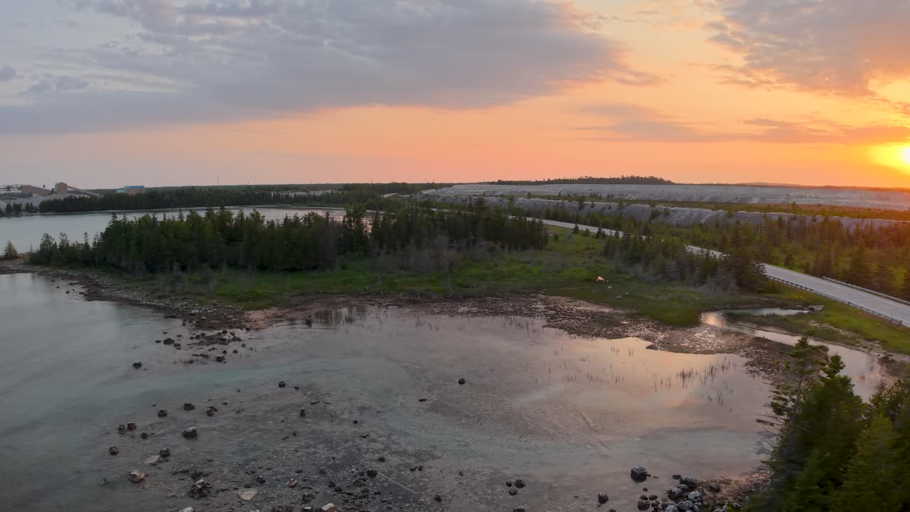 Aerial view of a coastal inlet with scattered rocks, forested shoreline, and a golden sunset over calm waters