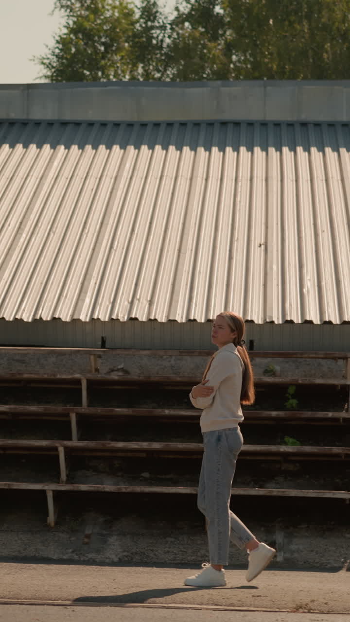 dama con capucha y vaqueros con el cabello largo atado hacia atrás caminando cerca de asientos vacíos del estadio en un día soleado, manos cruzadas, mirando reflexiva e introspectiva, rodeada de gradas vacías y árboles verdes