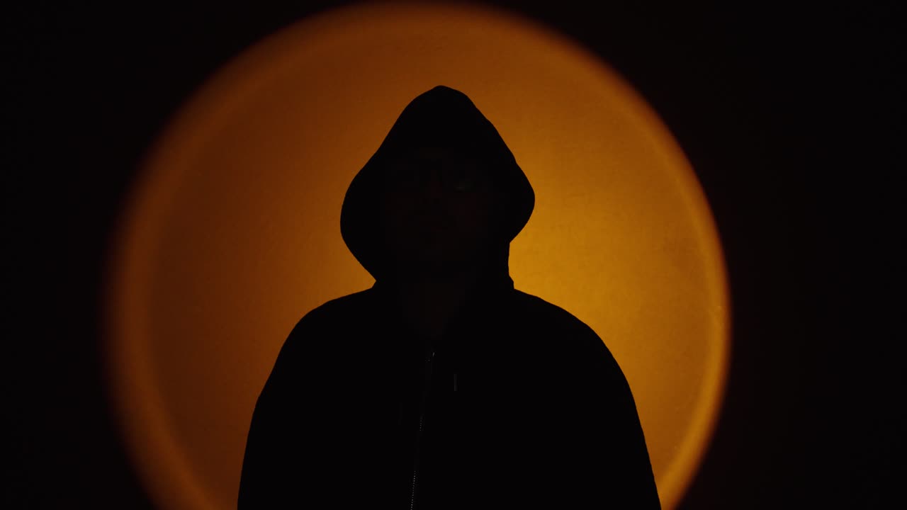 Silueta de un hombre anónimo con capucha, de pie contra una retroiluminación naranja circular. Atmósfera oscura y cinematográfica que transmite misterio, secretismo y anonimato