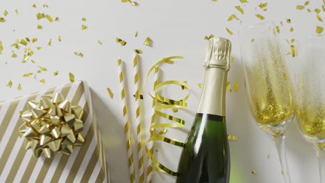 Video-of-champagne-bottle,-glasses-and-gifts-with-gold-streamers-and-confetti-on-white