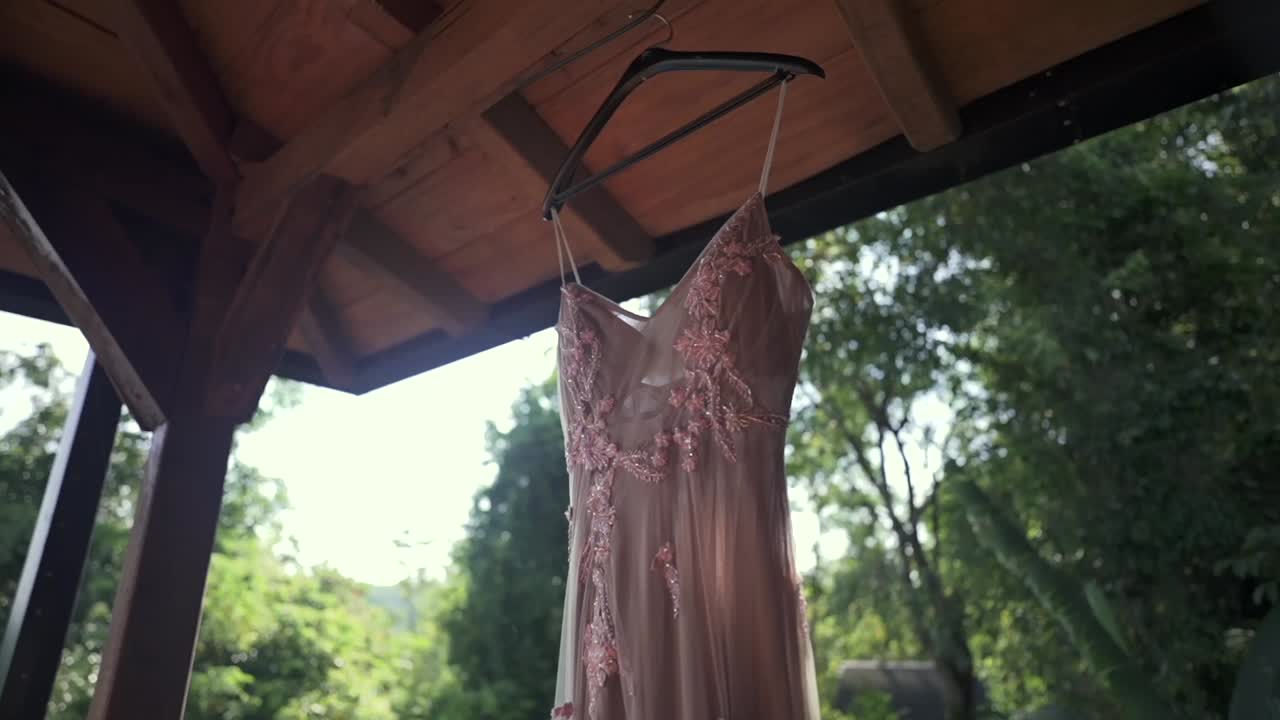 Elegant pink wedding dress hanging outdoors under wooden roof gazebo surrounded by green forest