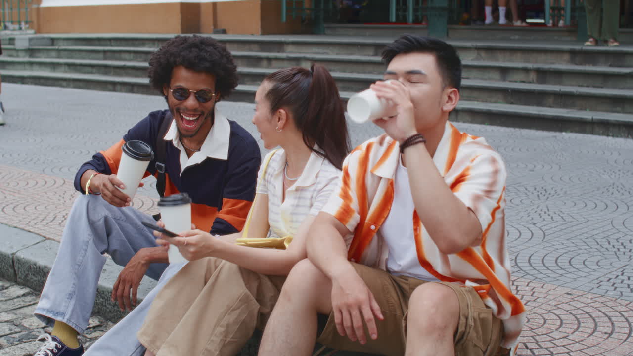 Friends Sitting on Pavement, Looking at Phone and Drinking Coffee at City