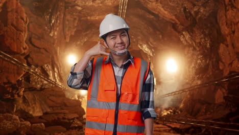 asian male engineer with safety helmet smiling to camera and making call me gesture while standing in underground mine tunnel