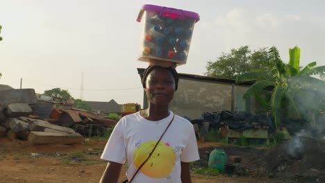 young woman with a smile carrying a plastic box on her head that looks like some bottles of drinks to be peddled at a community in kumasi, ghana