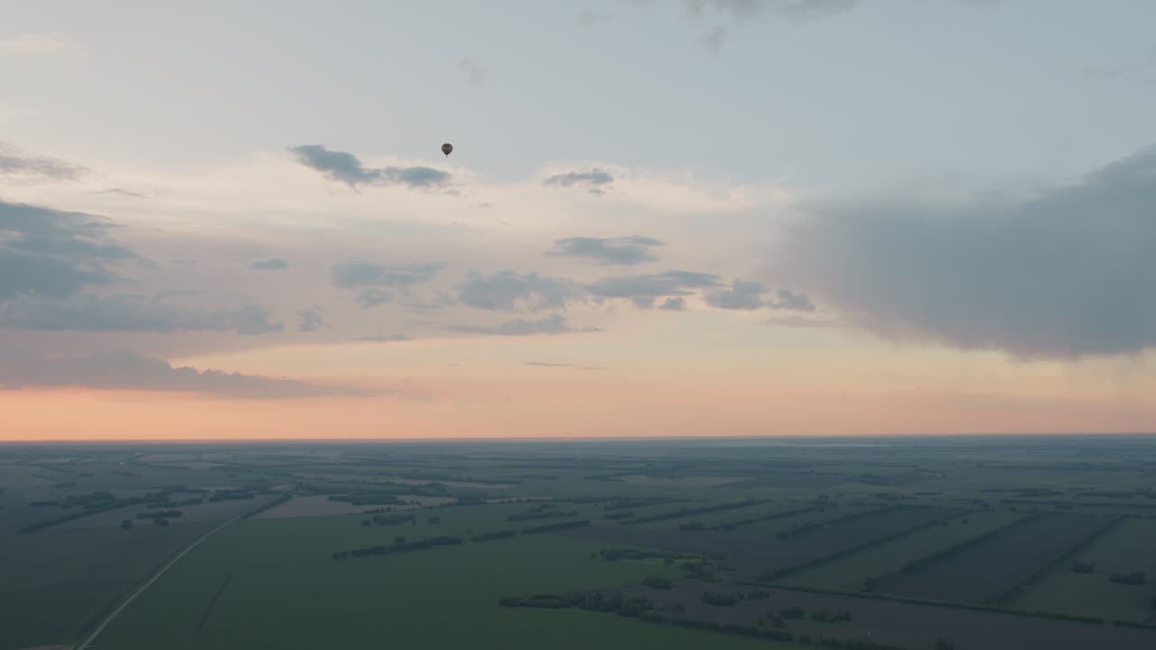 solitary hot air balloon drifts above patchwork farmland under pastel sunset sky offering serene aerial vista of green and brown fields with distant horizon and wispy clouds in warm evening glow