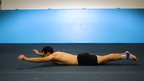 a guy doing an abs workout in a gymnastics gym doing lying down shortless leg and hand raises still shot