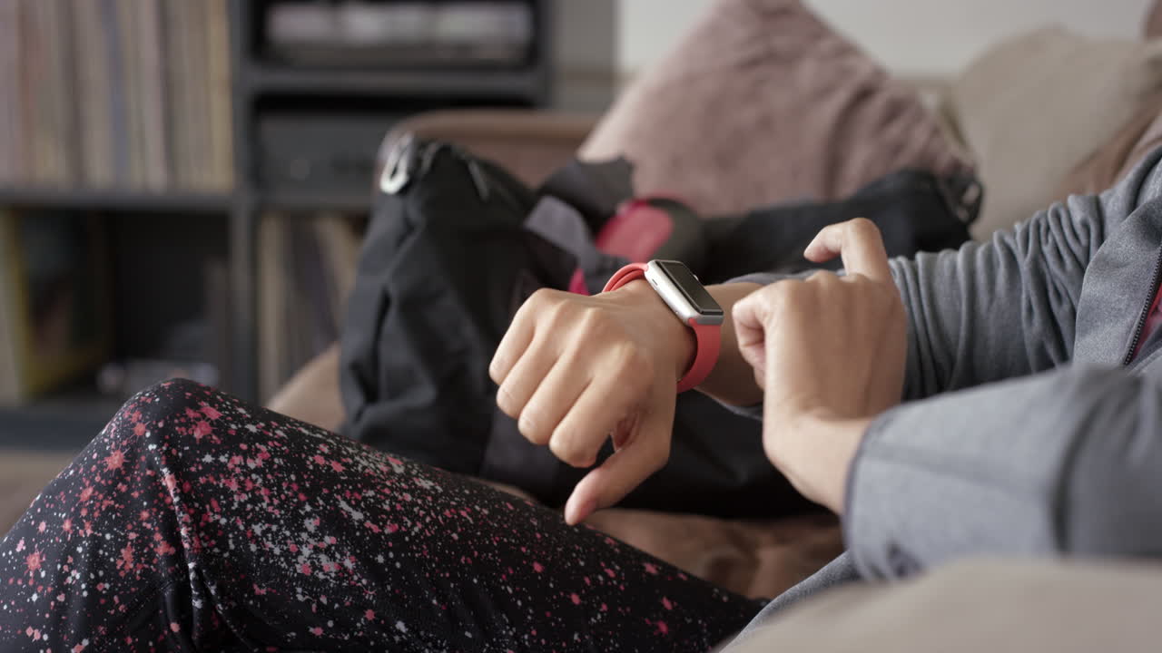Donna sportiva che, prima della palestra, controlla i messaggi sullo smartwatch a casa, rilassandosi sul divano.