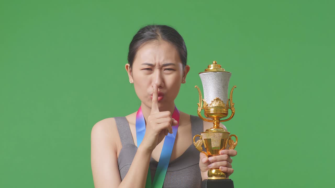 금메달과 트로피를 들고 있는 아시아 여성의 클로즈업, 한 제스처를 하고, '이것은 비밀이다'라고 말하며, 스튜디오의 녹색 스크린 배경에서 첫 번째 우승자로 승리합니다.
