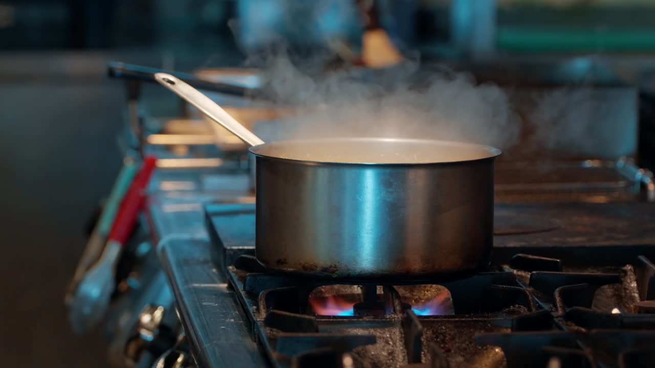 Close-up of a pot of soup simmering and steaming on a gas stove, capturing the bustling atmosphere of a professional restaurant kitchen as a chef passes by in the background