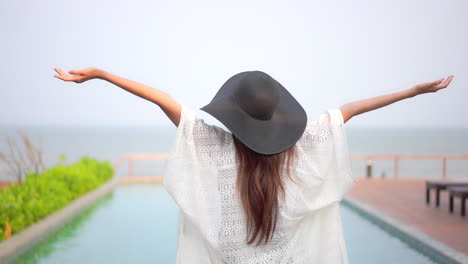 Espalda-De-Una-Mujer-Asiática-Parada-Junto-A-La-Piscina-Infinita-En-Verano-Usando-Un-Vestido-De-Verano-Y-Un-Sombrero-Negro-Levantando-Las-Manos,-Plantilla-De-Vacaciones