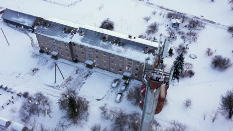 Small-apartment-building-and-tall-tele-communication-tower-in-winter-season-during-snowfall,-aerial-ascend-shot