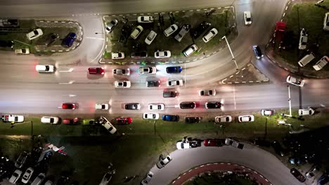 cars stop at traffic light rounding bend at night with vehicles parked on grass, aerial