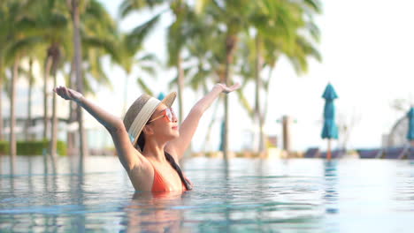 Mujer-Asiática-En-Traje-De-Baño-Naranja-Levantando-Los-Brazos-Dentro-Del-Agua-De-La-Piscina-Sobre-Fondo-De-Palmeras-Tropicales-Exuberantes-En-Bali-Al-Atardecer,-En-Cámara-Lenta